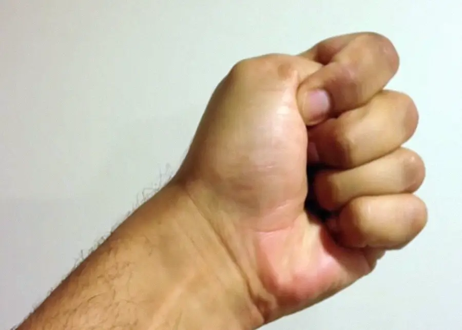 A close-up of a person's hand forming a fist, with the thumb resting on top of the curled fingers. The background is plain and light-colored.