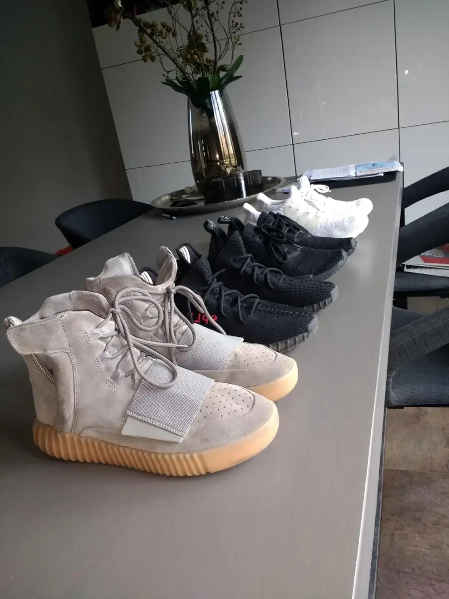 Five pairs of sneakers are lined up on a dining table, including beige high-tops at the front and several pairs of black and white low-tops behind them. Chairs and a vase with flowers are in the background.