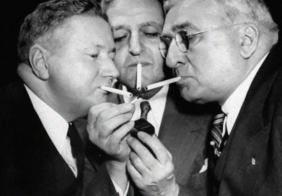 Three men in suits stand close together, each holding a cigarette to a lighter as they simultaneously try to light them, focused on the flame. The image is black and white and has a vintage feel.