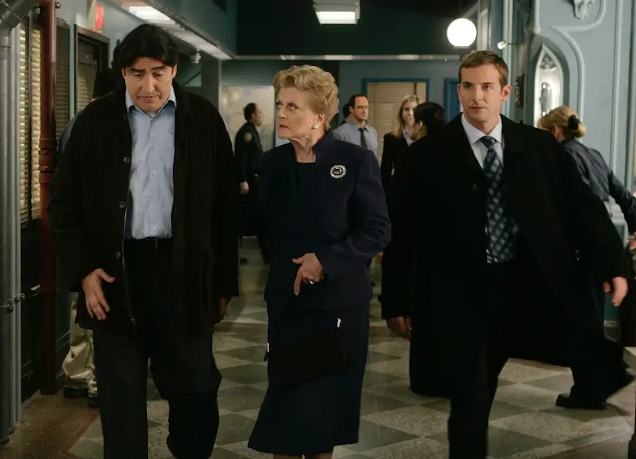 Three people, two men in dark coats and a woman in a navy outfit, walk together in a busy hallway with police officers and other people in the background. The setting appears to be a police station or official building.