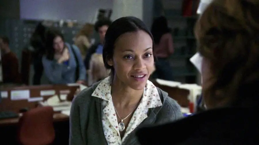 A woman with a light floral blouse, gray sweater, and necklace smiles while talking to someone in an office setting, with desks, papers, and other people blurred in the background.