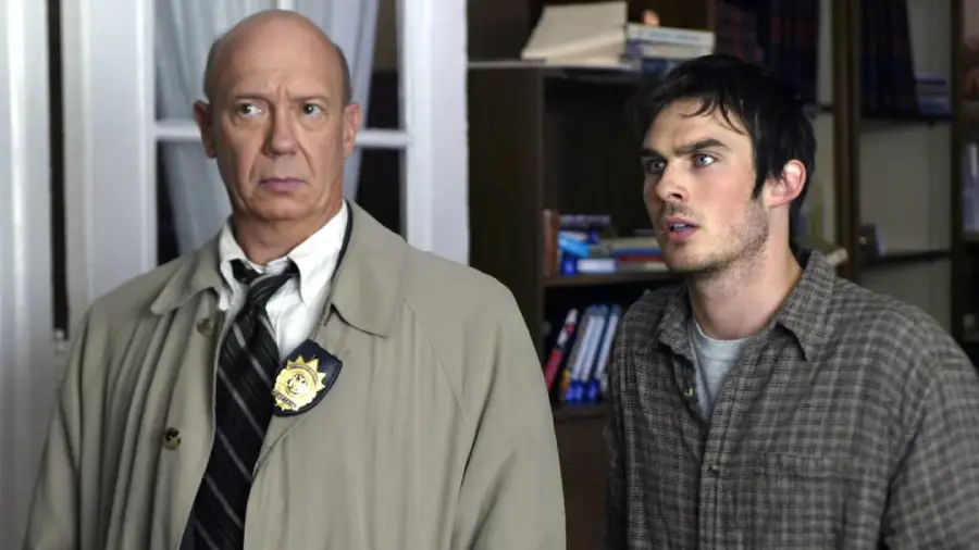 A concerned older man in a beige trench coat and police badge stands beside a younger man in a plaid shirt, both looking intently ahead in a room filled with bookshelves.