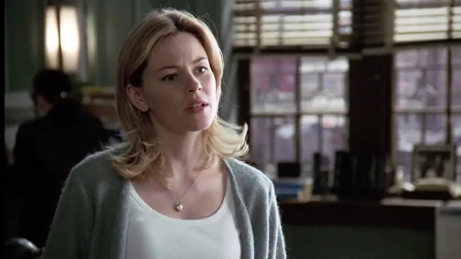 A woman with shoulder-length blonde hair, wearing a white top and gray cardigan, stands indoors in front of large windows with blinds, looking slightly to the side with a neutral expression.