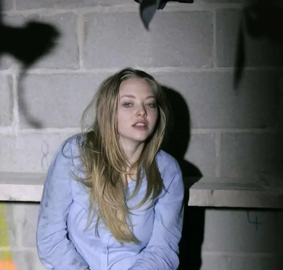 A woman with long blonde hair wearing a light blue shirt sits in front of a gray cinder block wall, looking slightly up with a relaxed expression. Shadows and soft lighting create a moody atmosphere.