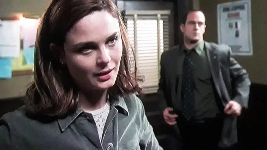 A woman with brown hair in a collared shirt stands in the foreground, looking serious, while a man in a suit and glasses stands behind her in an office with bulletin boards and blinds.