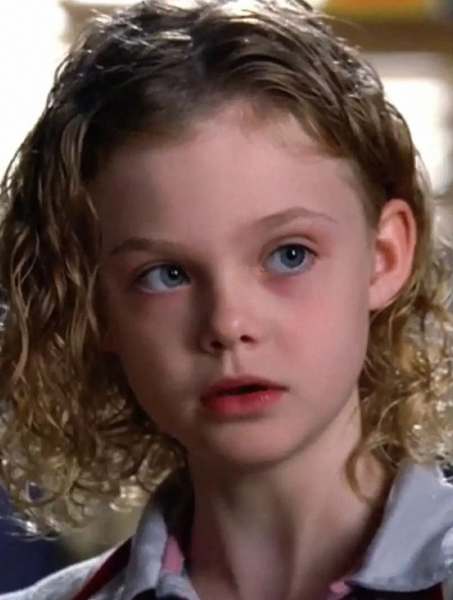 A young child with curly blonde hair and blue eyes looks slightly to the side with a curious and attentive expression. The background is softly blurred.