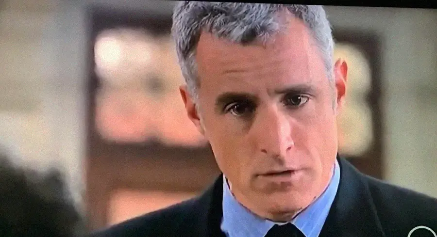 A middle-aged man with short gray hair, wearing a dark suit and blue shirt, looks slightly downward with a serious expression indoors. The background is blurred.