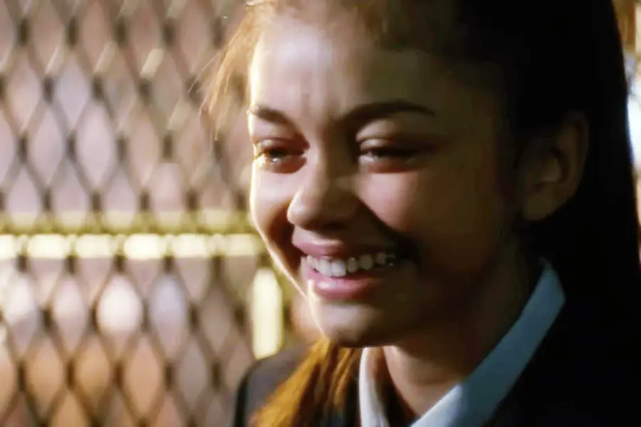 A young girl with long brown hair smiles brightly, showing her teeth. She is wearing a dark jacket and is standing in front of a metal fence or grate, with warm light in the background.