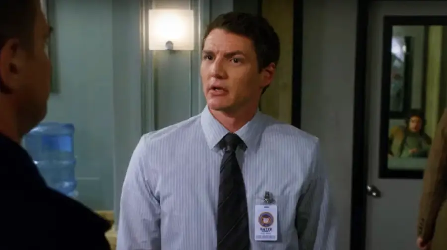 A man in a striped shirt, tie, and ID badge stands indoors, speaking to someone off-camera. A water cooler and window are visible in the background.