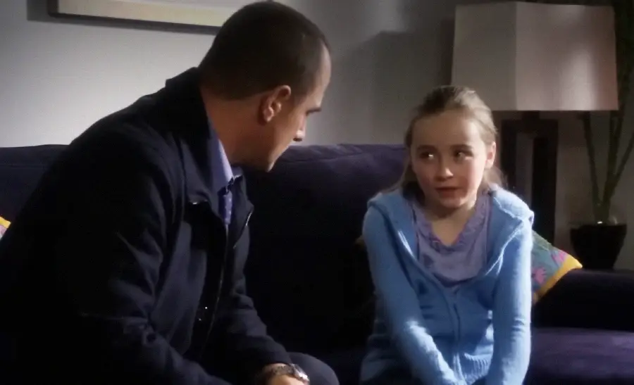 A man in a dark jacket sits on a couch next to a young girl in a blue hoodie. The man looks at her attentively while she looks away, appearing uncomfortable. They are indoors with a lamp and plant in the background.