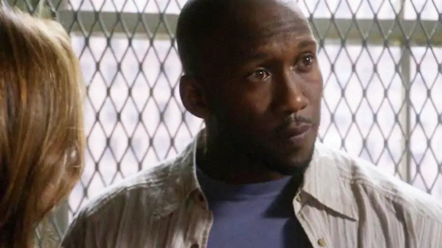 A man with a shaved head and trimmed beard, wearing a light-colored shirt over a blue t-shirt, stands indoors in front of a window with a metal grid, looking thoughtfully to the side.