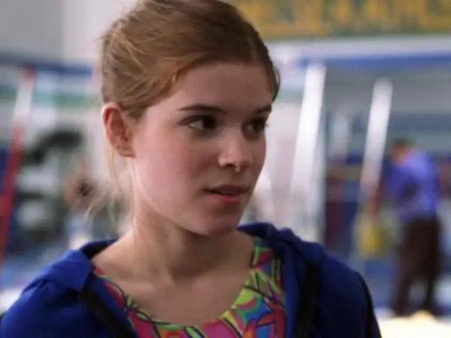 A young woman with light skin and auburn hair pulled back is wearing a colorful patterned top and a blue jacket. She is indoors, possibly in a gym, and looks slightly to the side with a neutral expression.