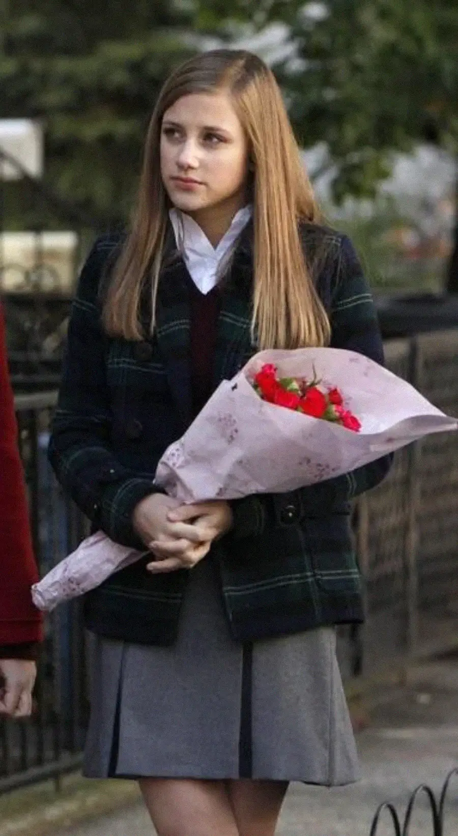 A young woman with straight, light brown hair stands outside holding a bouquet of red roses wrapped in pink paper. She wears a plaid jacket, white shirt, and gray pleated skirt, looking thoughtful. Trees and a fence are in the background.