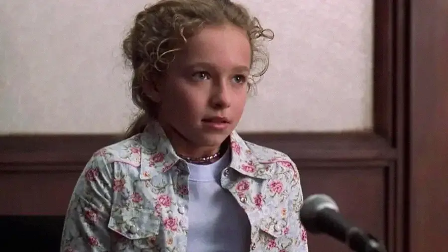 A young girl with curly blonde hair, wearing a white shirt and a floral jacket, sits in front of a microphone in what appears to be a courtroom or formal setting, looking slightly nervous or uncertain.