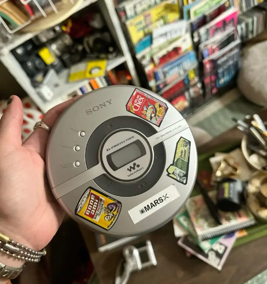 A hand holds a silver Sony portable CD player decorated with colorful stickers. In the background are shelves filled with books and CDs, and a cluttered table with various items.