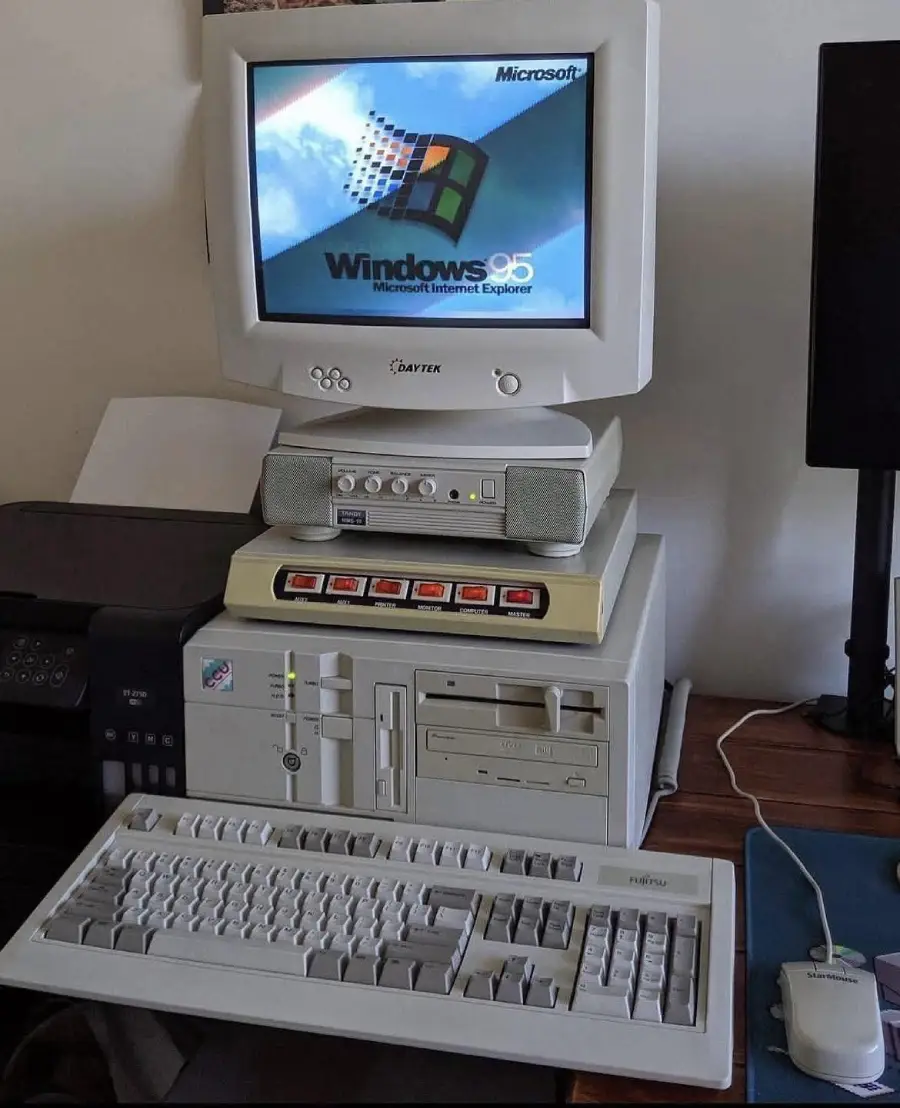 A vintage computer setup with a CRT monitor displaying the Windows 95 startup screen, a beige tower with floppy and CD drives, external speakers, a keyboard, and a wired mouse on a wooden desk.