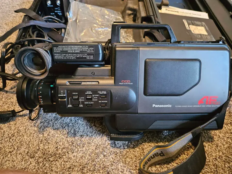A vintage Panasonic AF camera recorder with a lens, various control buttons, and a carrying strap, resting on a carpeted floor next to its open case and accessories.
