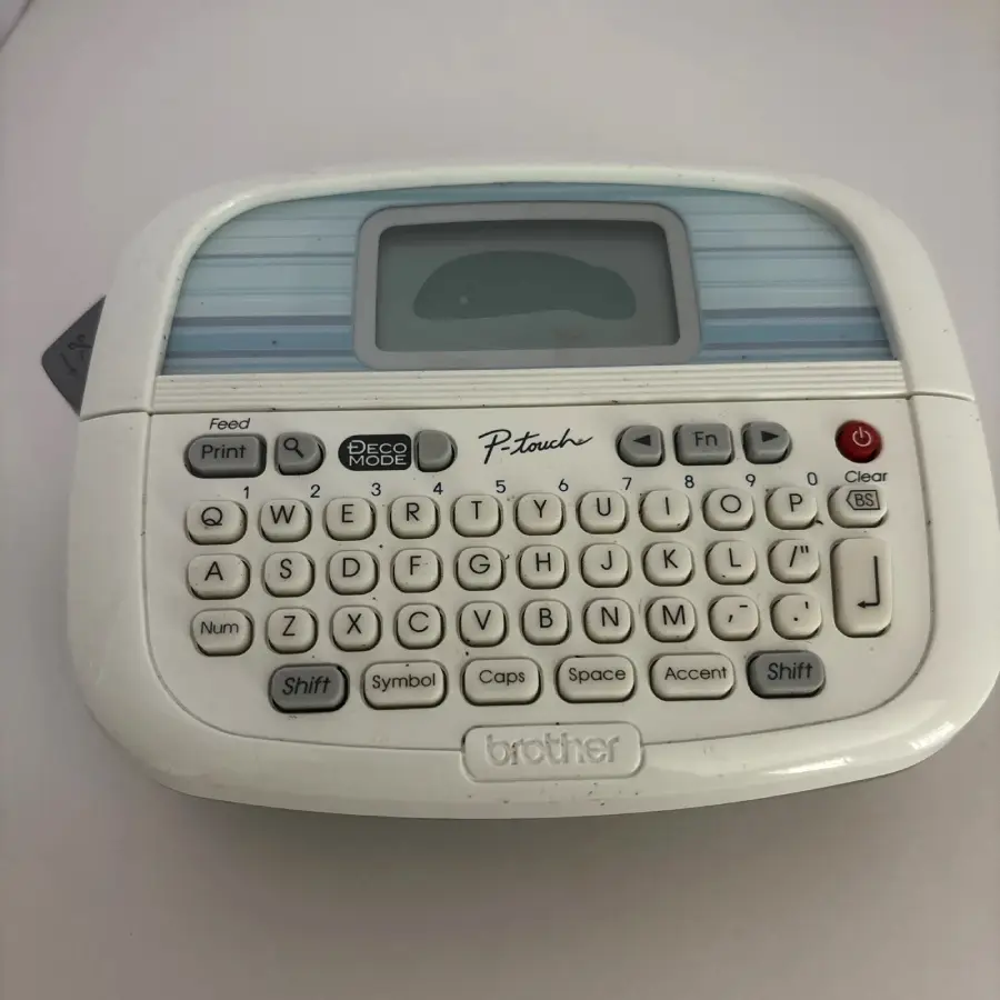 A Brother P-touch label maker with a QWERTY keyboard, small screen, and various function keys, set on a white surface.