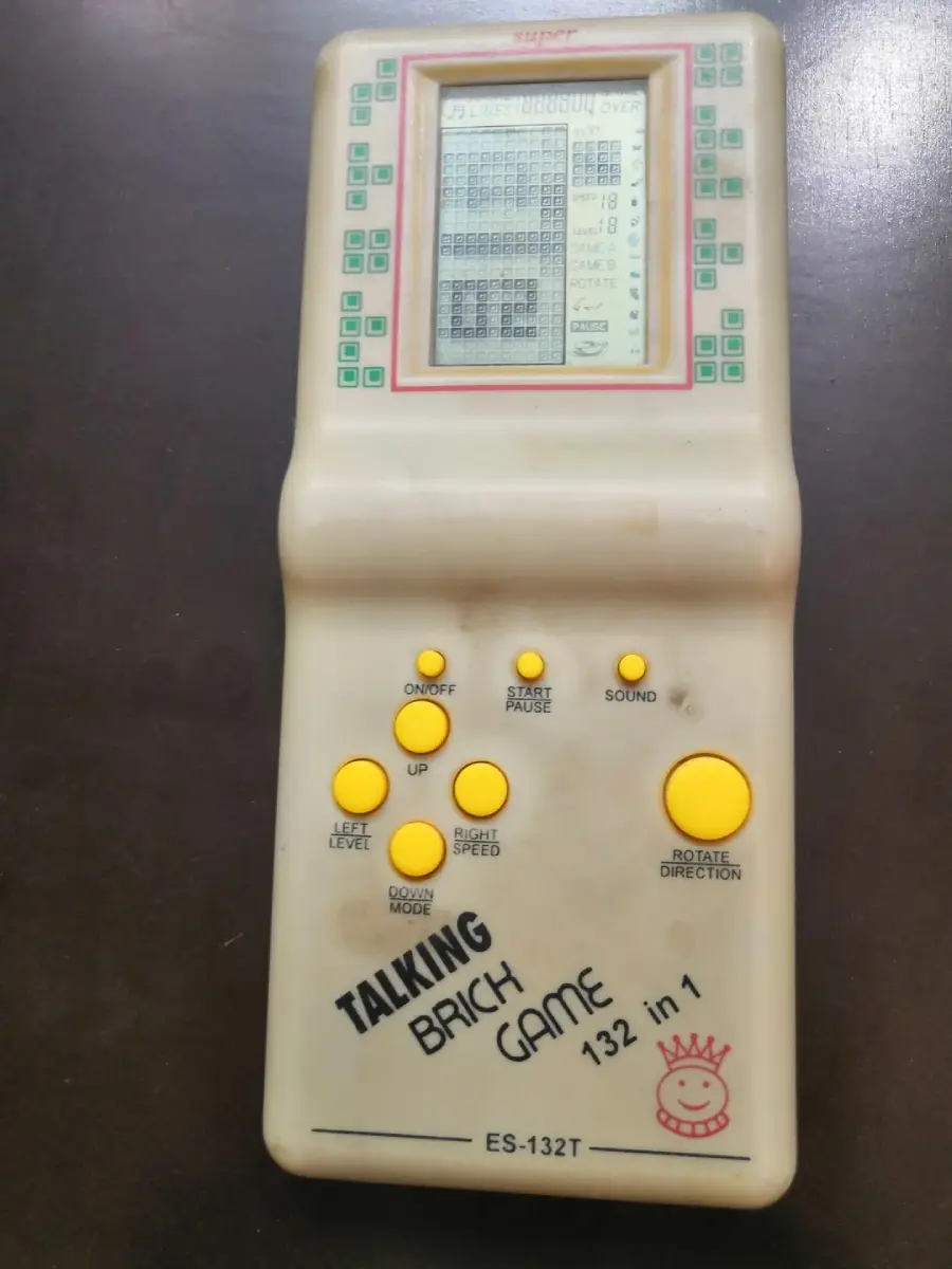 A vintage handheld "Talking Brick Game" console with a small monochrome screen, yellow control buttons, and the label "132 in 1" on a cream-colored plastic body.