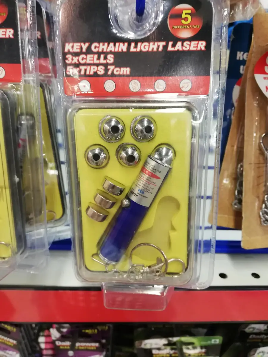 A packaged keychain light laser set with a blue laser pointer, batteries, and multiple interchangeable tips, displayed on a store shelf. The package advertises 3 cells and 5 laser tips.