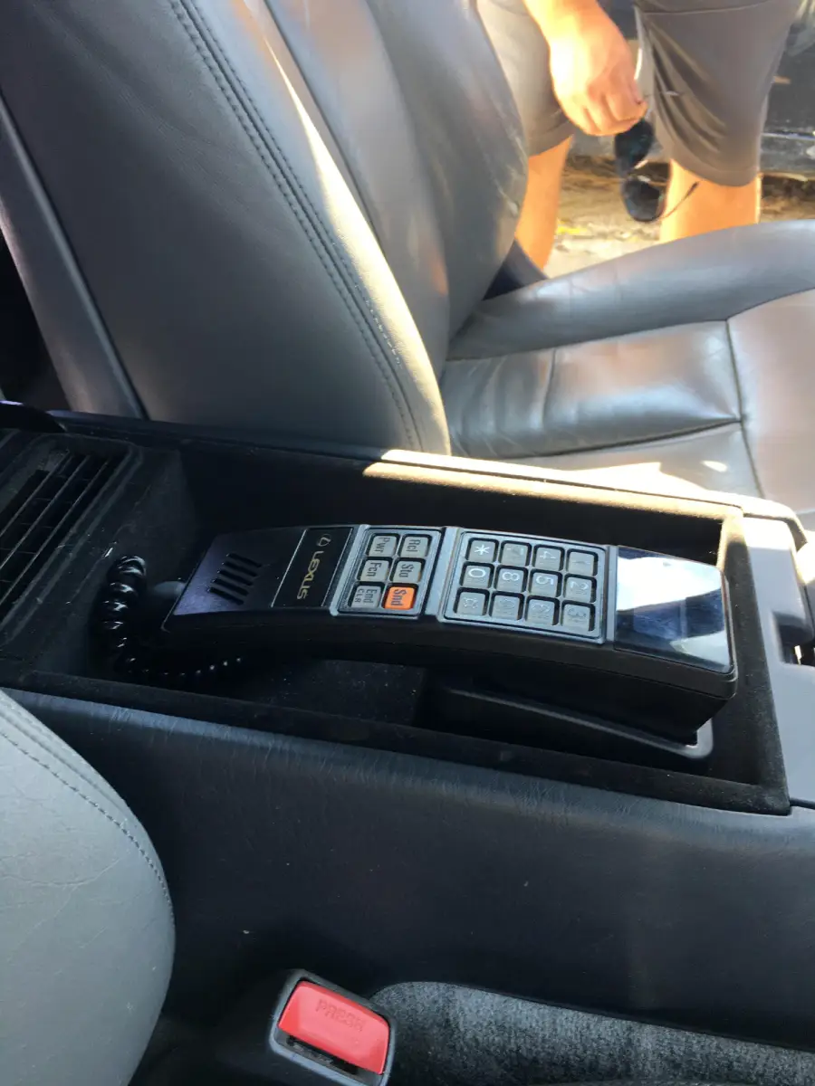 A vintage car phone with a cord is installed in the center console of a car with leather seats. A person’s legs are visible outside the vehicle near the open door.