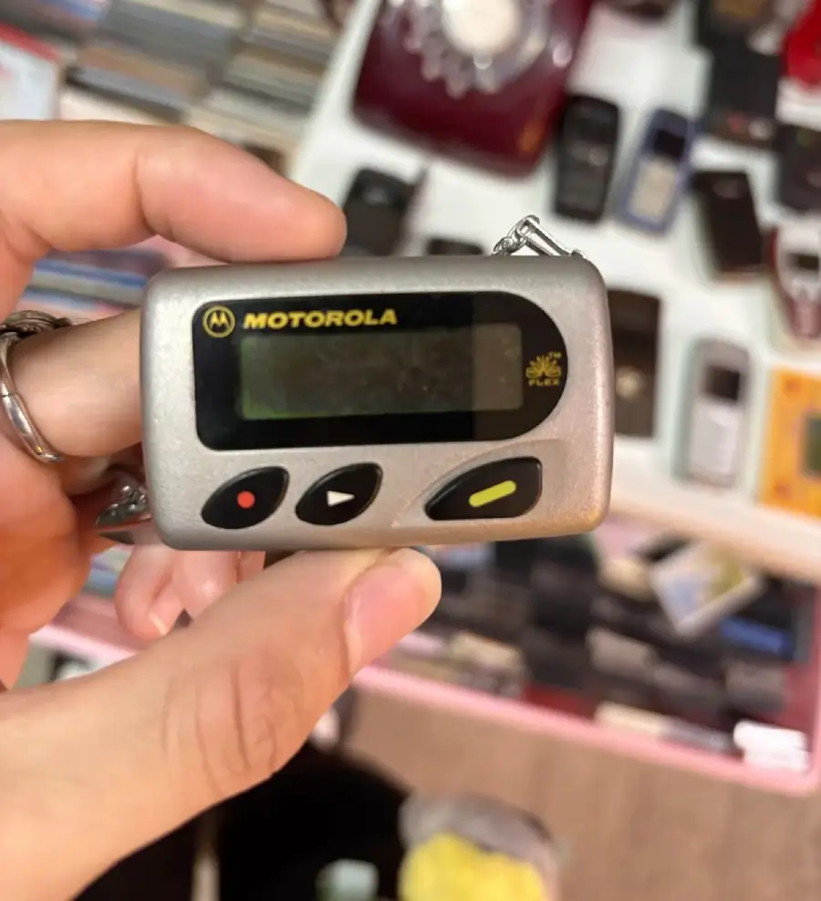 A hand holds a Motorola pager with three buttons, in front of a blurred background displaying various old electronic devices.