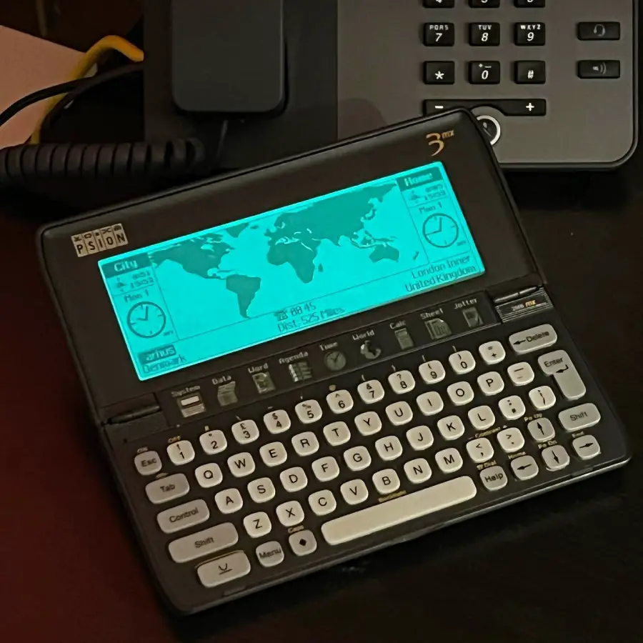 A Psion Series 3mx handheld computer displays a world clock and map on its screen, showing different time zones. The device has a small QWERTY keyboard and is placed on a desk near a black office phone.