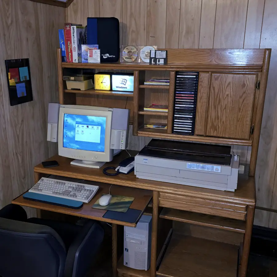 A vintage computer desk setup with a CRT monitor displaying Windows, keyboard, speakers, an old printer, floppy disks, CD cases, office supplies, and wood-paneled walls in a small, organized workspace.