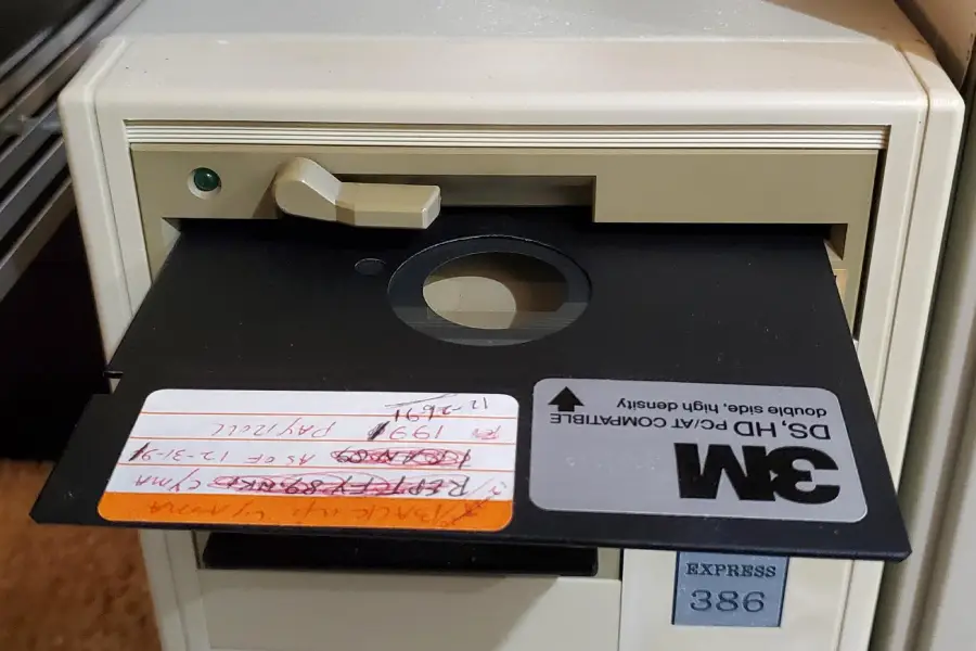 A close-up of an old computer with a 5.25-inch 3M floppy disk partially inserted into its drive. The disk has handwritten labels and the drive is labeled "RXPR888 386.