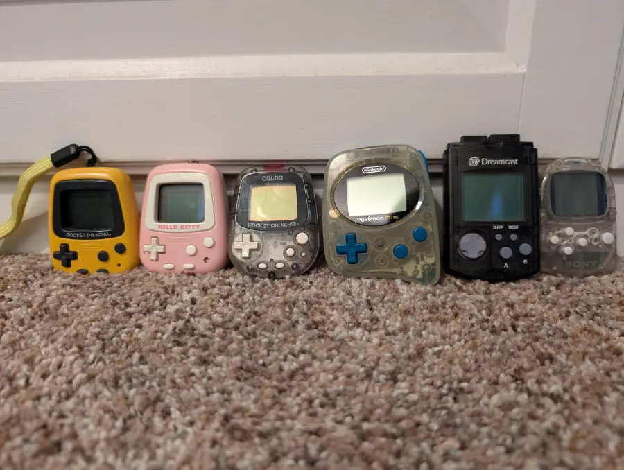 Six handheld virtual pet and gaming devices, including a yellow, pink, gray, green, black, and clear unit, are lined up side by side on a beige carpet in front of a white door.