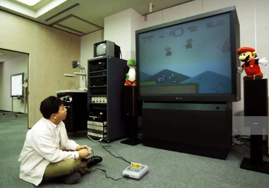 A person sits on the floor playing a Super Nintendo game on a large TV in an office. Plush Yoshi and Mario toys are on top of the TV, with audio-video equipment nearby.