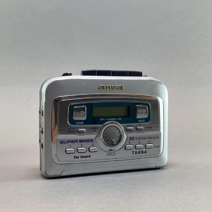 A silver Aiwa TX494 portable radio with buttons and a digital display is shown against a plain gray background. The front panel features station presets, tuning controls, and "Super Bass" branding.