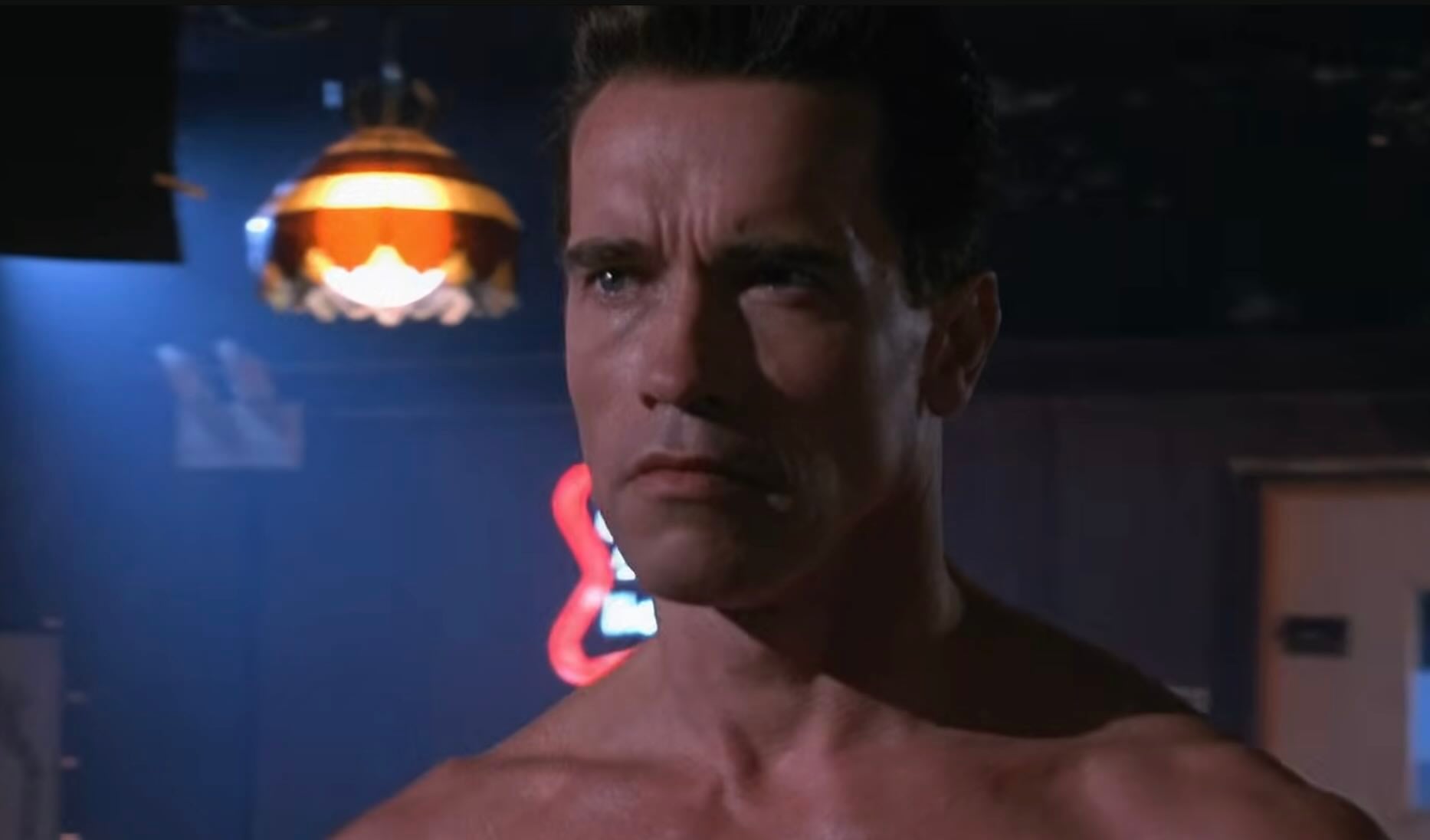 A muscular man with a serious expression stands shirtless in a dimly lit bar, illuminated by overhead lights and neon signs in the background.