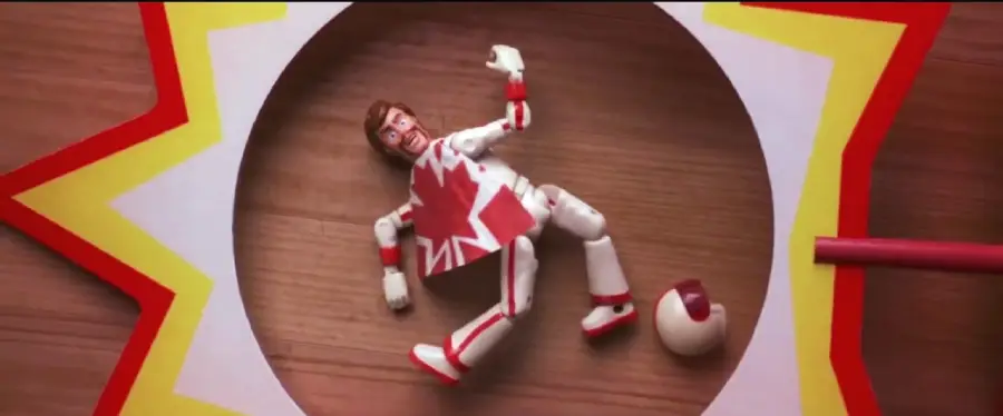 A toy action figure in a red and white outfit with a maple leaf lies awkwardly on a wooden surface next to a small ball, inside a circular frame with a jagged yellow and red border.