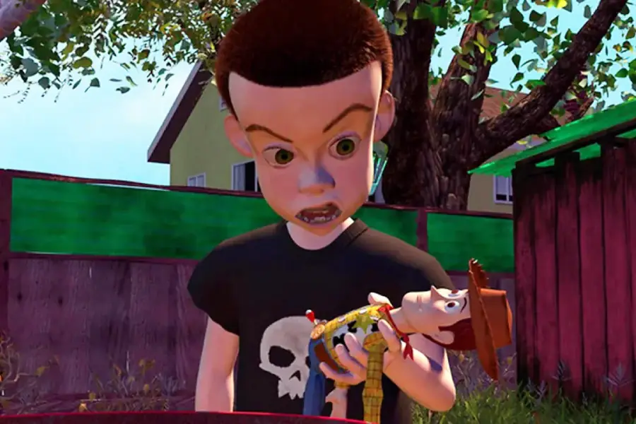 Animated boy with a black shirt featuring a white skull glares menacingly at a cowboy doll he’s holding outdoors, with trees, a wooden fence, and houses in the background.