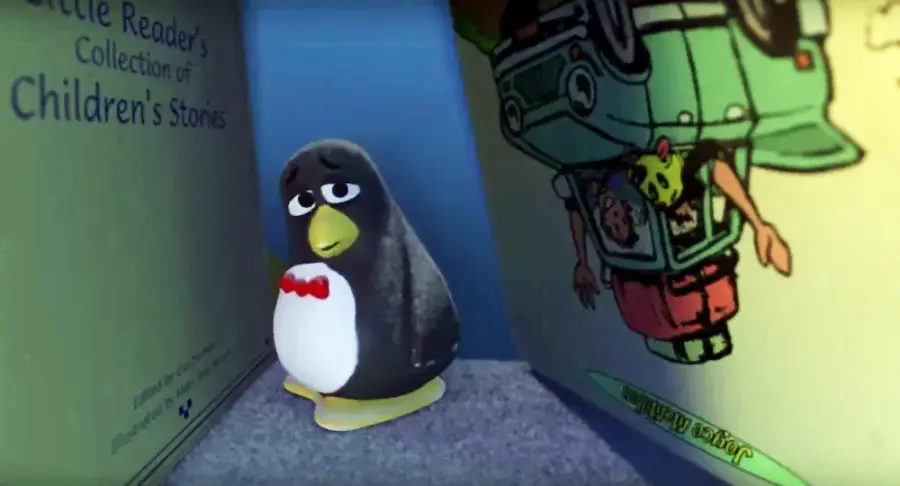 A stuffed penguin toy with a red bow tie stands between two oversized children’s storybooks, one titled "Little Reader’s Collection of Children’s Stories." The penguin appears to be looking up sadly.