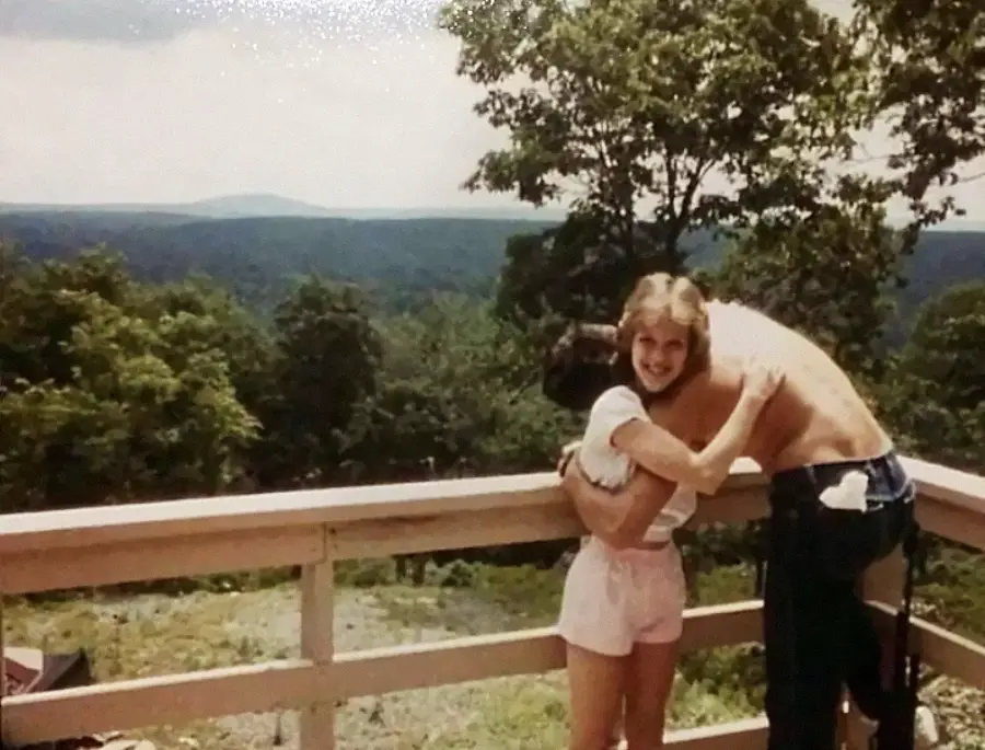 Two people hug on a wooden deck overlooking a forested landscape. One person is shirtless, the other wears a white shirt and shorts. The background shows trees and distant hills under a partly cloudy sky.