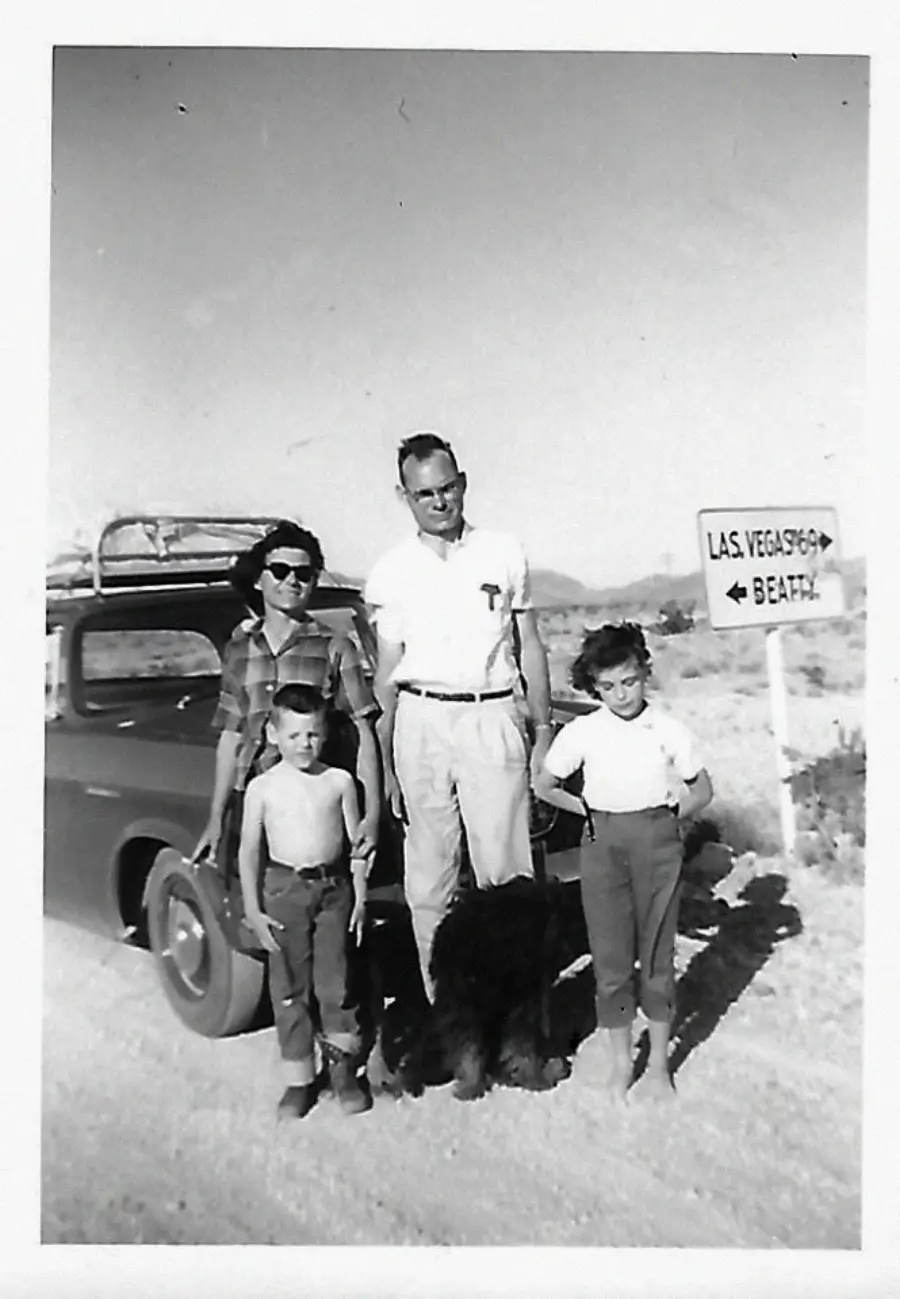 A black-and-white photo of a family with two children and a dog standing beside a vintage car on a dirt road. A road sign points to Las Vegas and a beach. Desert landscape in the background.