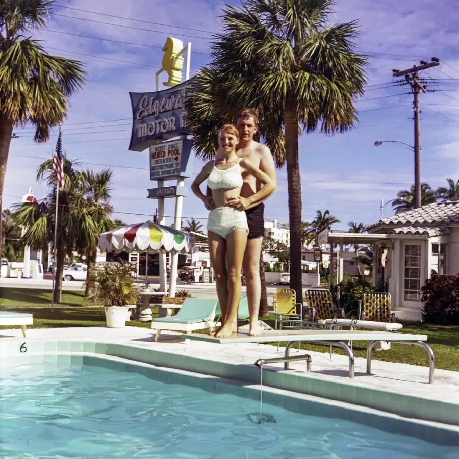 A smiling couple in vintage swimwear stands beside a pool at a mid-century motel, surrounded by palm trees. A retro sign reading "Laguna Motor" is visible in the background under a sunny sky.