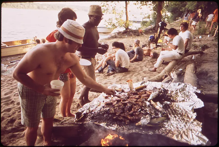 People gather on a sandy riverbank, cooking meat on a large grill covered with foil. Some stand and grill, while others sit, eat, and talk near the water, with trees and a boat visible in the background.