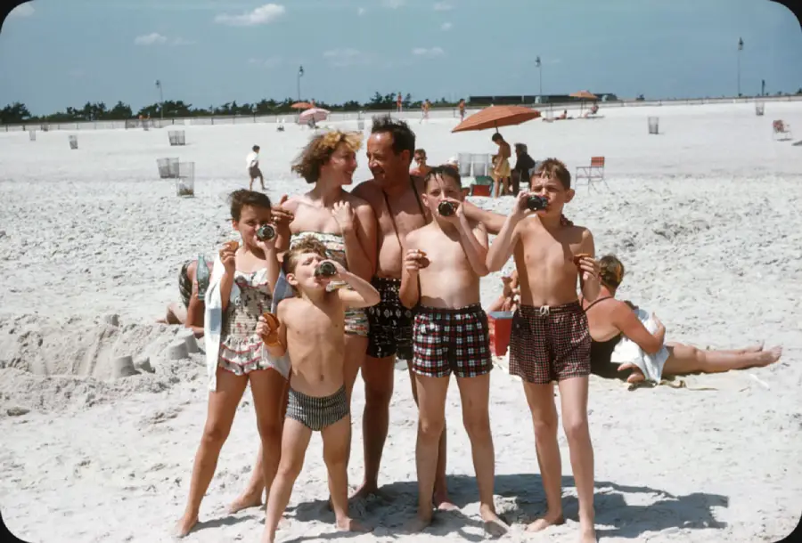A family of six poses on a sandy beach, all wearing swimsuits. Four children in front hold cameras, while two adults stand behind them. Other beachgoers and umbrellas are visible in the background.