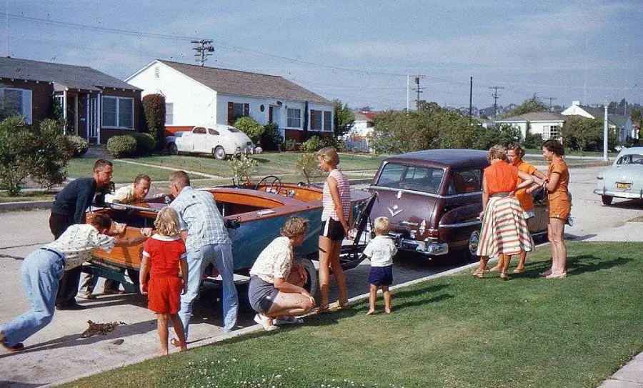 A group of adults and children gather around a car and a wooden boat on a suburban street, with 1950s-style houses and parked cars in the background. Some people are unloading or examining the boat.