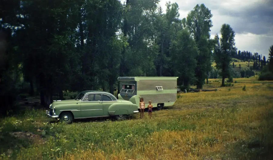 A vintage green car is parked in a grassy field, hitched to a small travel trailer. Two children stand nearby, surrounded by tall trees and wildflowers under a cloudy sky.