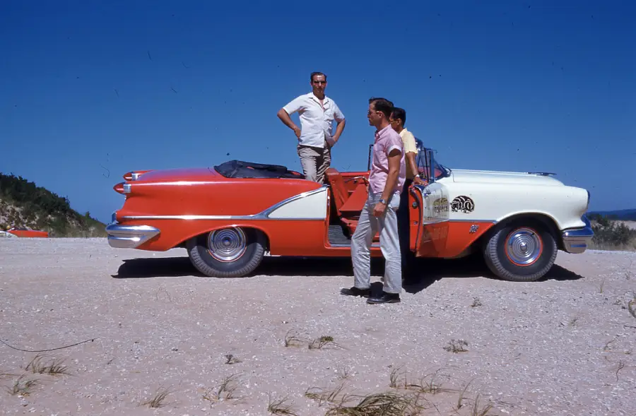 Three men stand next to a red and white vintage convertible car parked on a sandy area under a clear blue sky. One man stands inside the car, while the other two stand nearby, engaged in conversation.