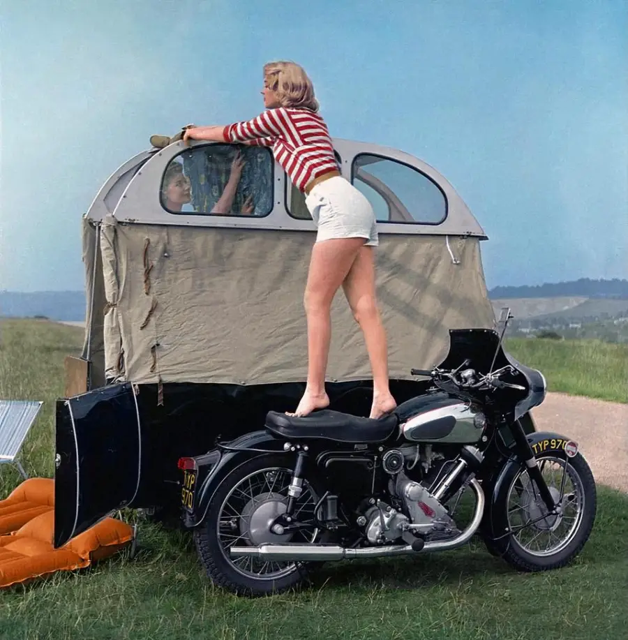 A woman in a striped shirt and white shorts stands on a motorcycle to reach into a vintage tent trailer, with a child looking out the window; the scene is set in a grassy countryside.
