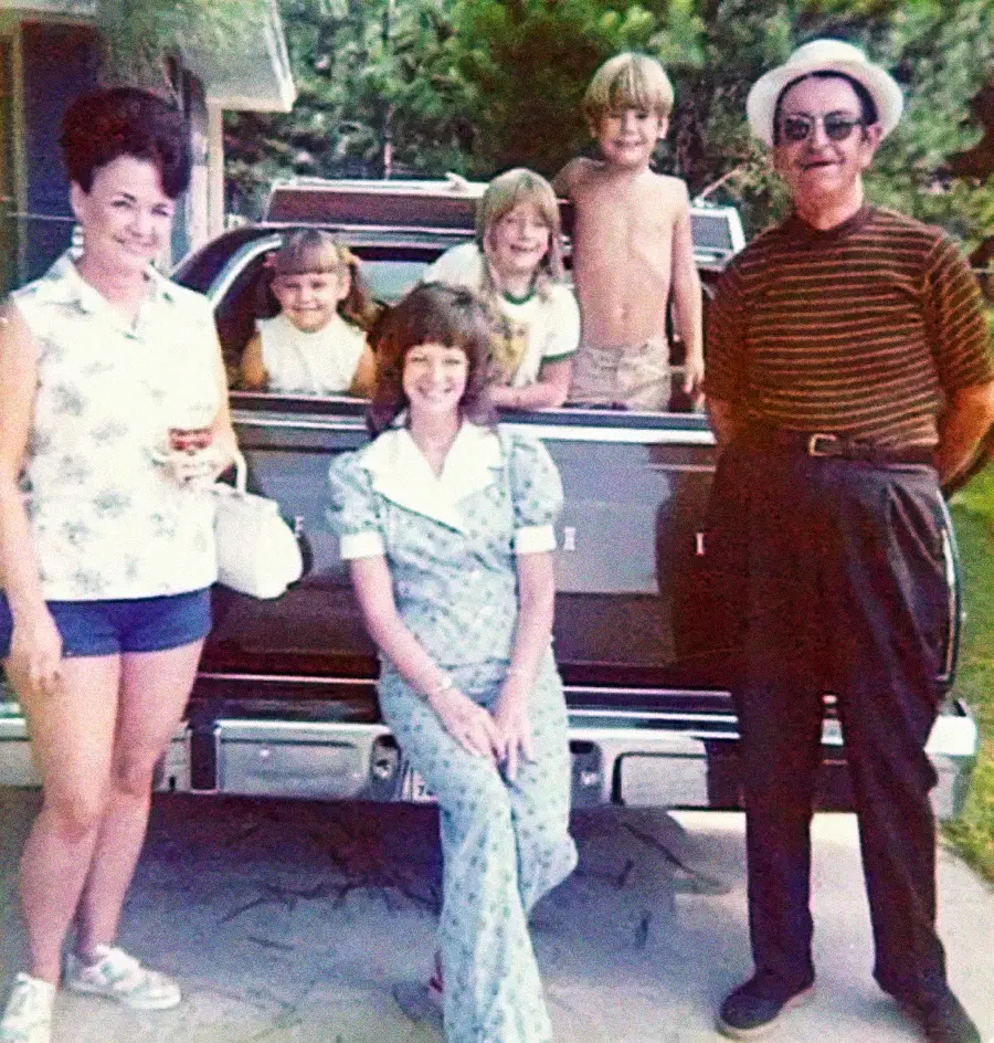 A vintage photo of a smiling family poses in front of a classic car. Two women and a man stand, while three children, one shirtless, lean over the car's trunk. Green trees and a house are visible in the background.