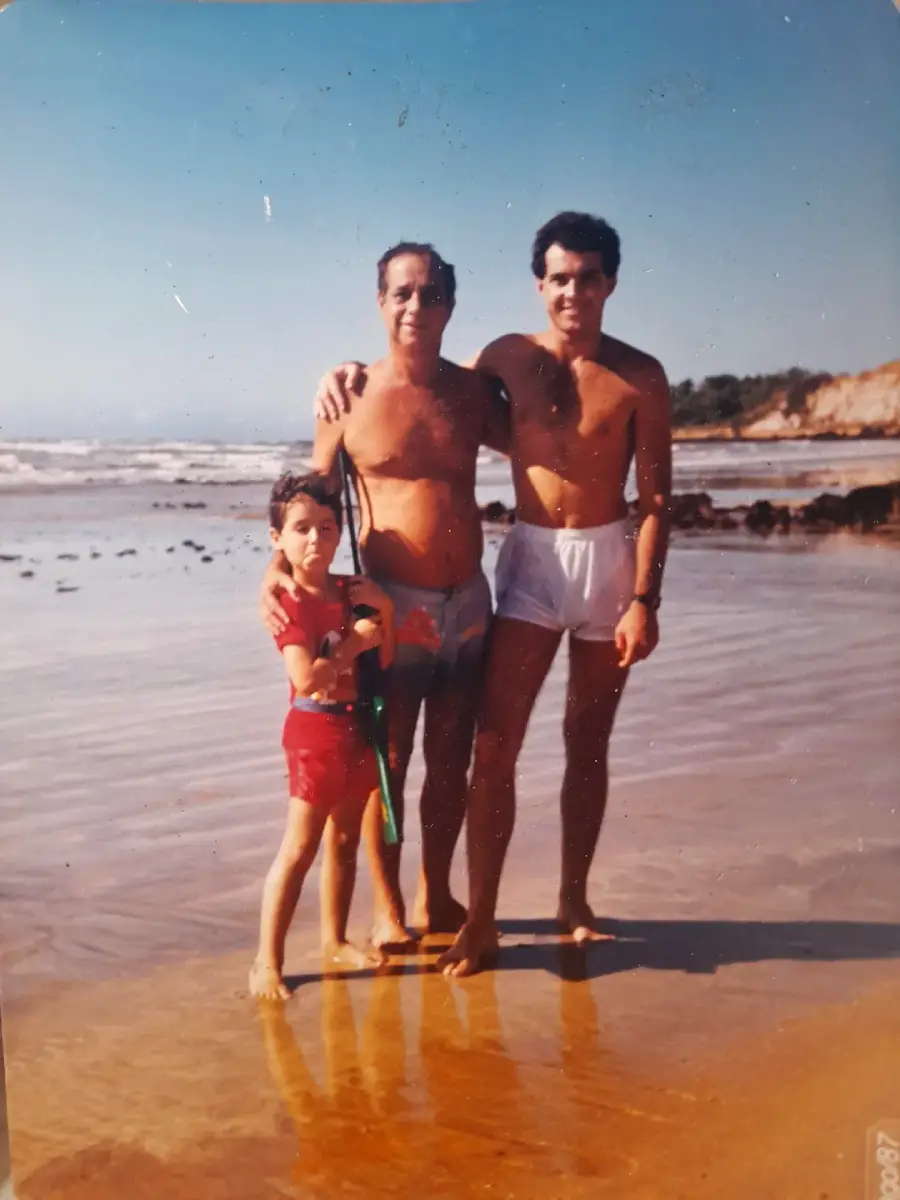 Three people stand on a sandy beach near the water: a child in red shorts holding a toy, and two adults in swim trunks with their arms around each other, all smiling on a sunny day. Cliffs and ocean are in the background.