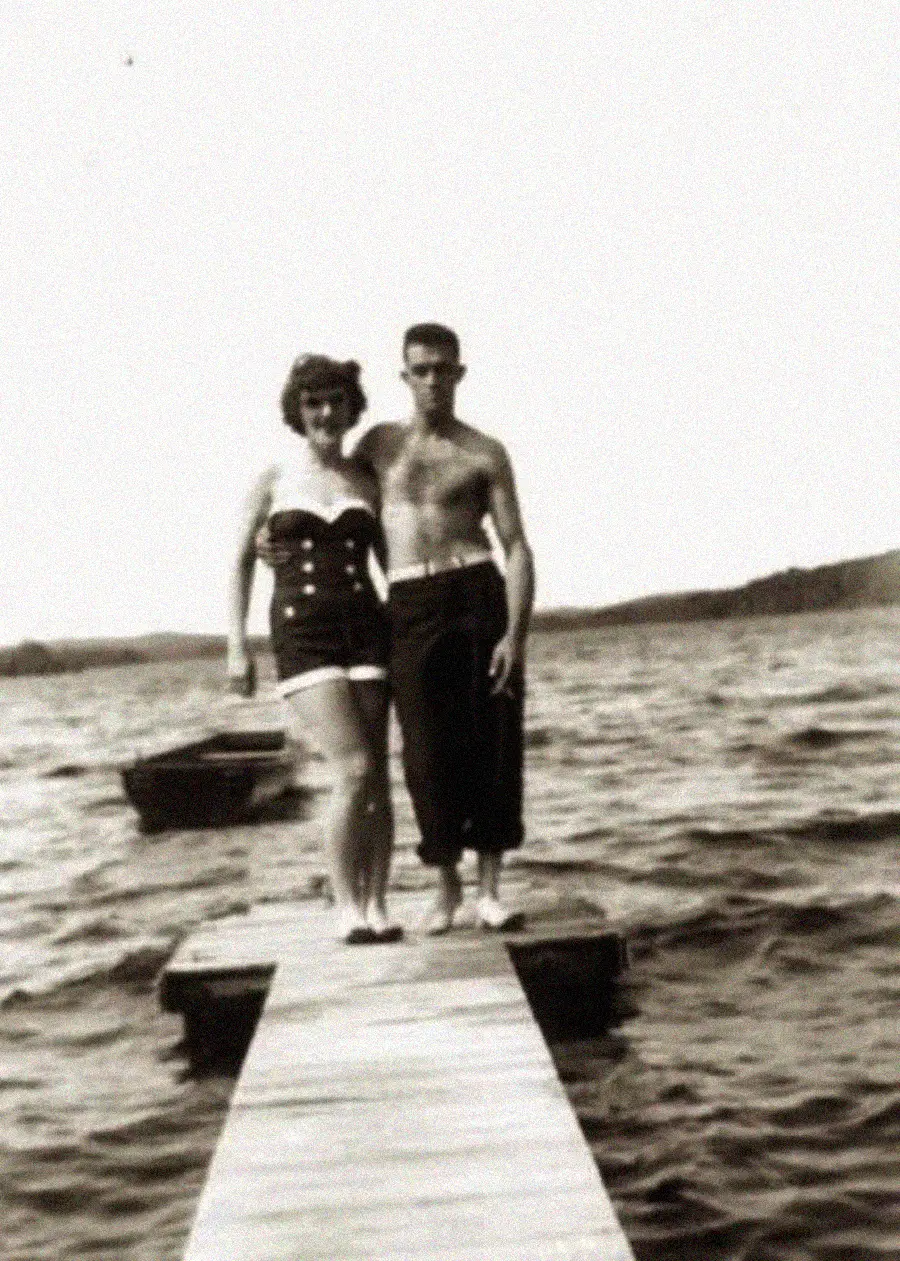 A vintage black and white photo shows a woman in a swimsuit and a shirtless man standing arm-in-arm on a wooden dock by a lake, with choppy water and a rowboat in the background.