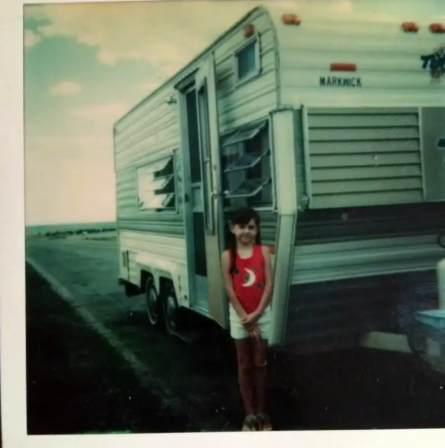 A young girl stands smiling in front of a parked travel trailer on the side of a road. The trailer is light-colored with silver trim, and the sky in the background looks overcast.
