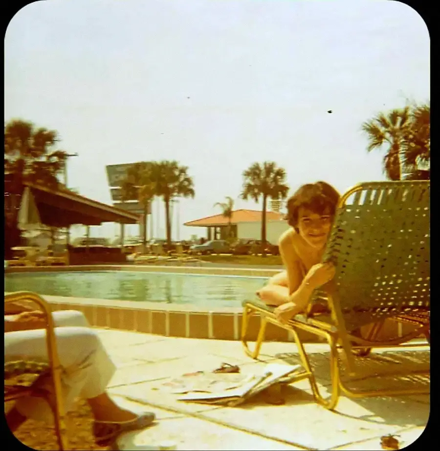 A person smiles while sitting on a lounge chair by a pool with palm trees in the background. Another person’s legs are visible on the left. It appears to be a sunny day, and there are buildings and a billboard in the distance.